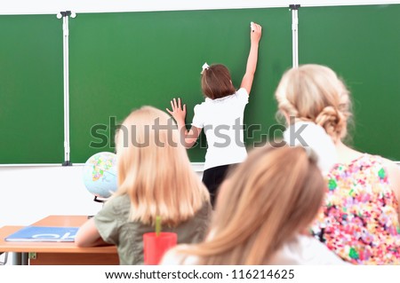school girl writes on the blackboard in class sitting other students and a teacher and look at the board