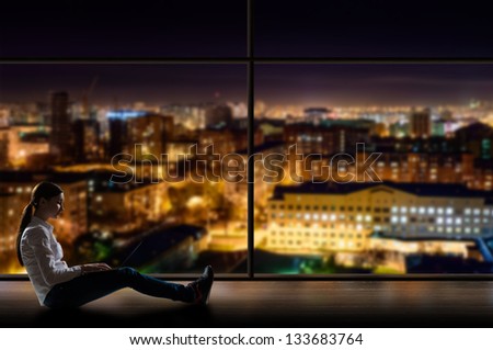 young woman sitting with a laptop by the window with a night city