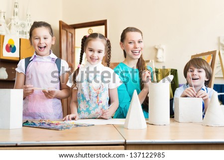 children with the teacher standing at the table, smiling and looking into the cam