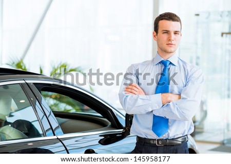 dealer stands near a new car in the showroom, folded his arms across his chest