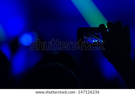 Silhouette of hands holding a mobile phone and taking a video of a rock concert. Low light photos, some grain present.