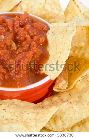 vertical closeup of chip in red bowl of hot salsa surrounded by salty snacks
