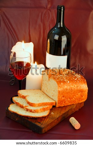 glass of red wine with dark colored bottle, lit candles and partially sliced loaf of bread on wooden board against burgundy background