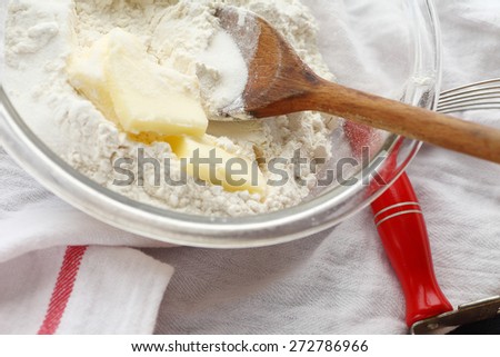 Glass mixing bowl with wooden spoon, butter and flour with pastry blender alongside on dish cloth
