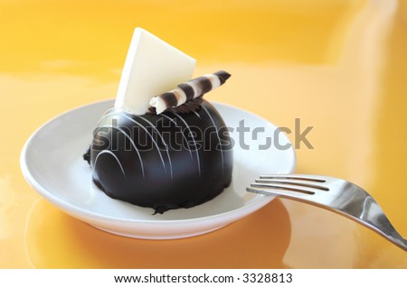 a chocolate dessert with a white chocolate decoration on a white plate with a fork against a reflective medium yellow background