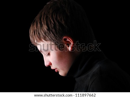 Teenage boy is photographed in profile on the black background. He is upset and his head are hung. She is wearing in black.