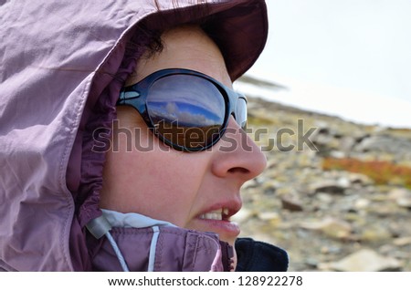 A face of hiker is photographed in profile on the blurred background of the slope with snowfield. The sky and mountains are reflected on her sunglasses.