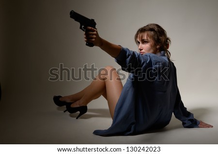 Young woman is sitting on the floor and aiming with gun. She is wearing a male shirt and high heels.