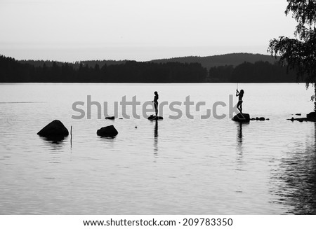 Teenage girl and boy are standing on stones and fishing rod in the big forest lake. They are photographed at sunset. Black-and-white.