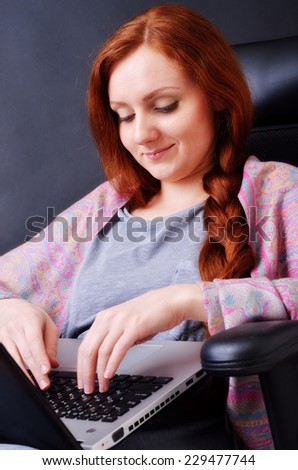 A happy young woman is sitting in the armchair at home. She types on a laptop and smiles.