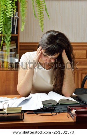 A young woman is reading a big book on the table. She enfolds her head both arms.