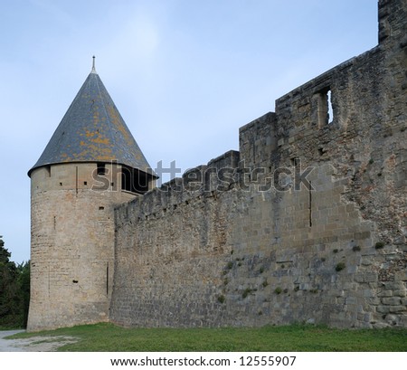 Old defense stone wall recovered with rough masonry and conic tower of Carcasson castle in sunlight against blue sky, France