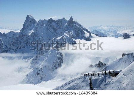 Mountain range above layer of white clouds, snow path with mountain-skiers in the foreground, Mont Blanc