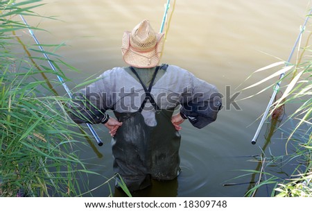 Fisherman stands in straw hat and wader akimbo before three rods, rear view, ripples in the still water