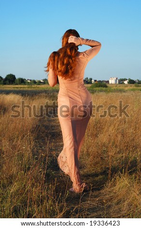 Long-haired girl going on path in transparent flesh-colored dress against rural landscape, hands hold red hair
