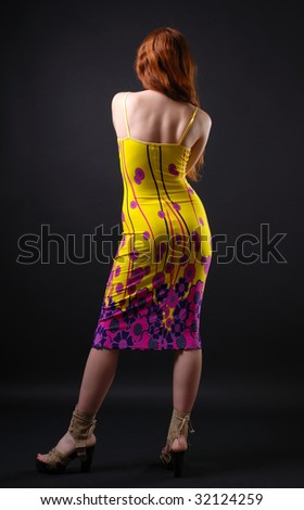 Redheaded girl standing back on dark background. Yellow close dress with shoulder-straps accentuate slender body. Knitted cloth of gown is ornamented with violet and rosy flowers.