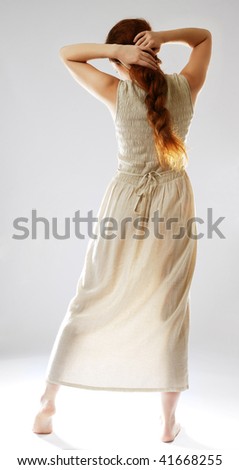 Young woman is standing back and tidying a heavy braid. She is wearing a long linen dress. Her maidenly body is illuminated from beneath. She is barefooted and isolated on white.