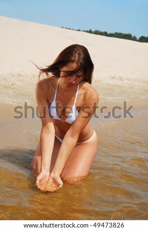 Pretty girl is sitting in the shallow water and drawing water. In the background there is the sand beach. She is wearing a white bikini.