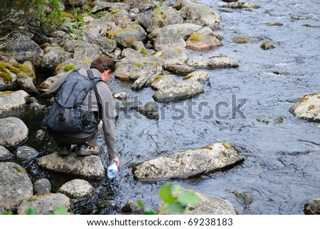 Young man is sitting on the stony riverside and filling the bottle with water.