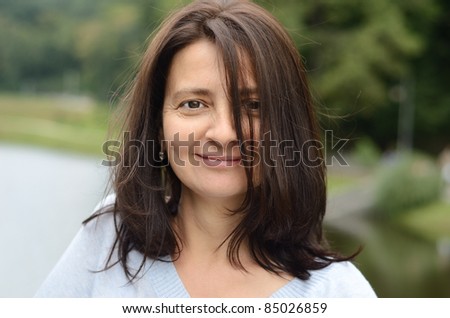 Pleased woman is photographed outdoors. There is the green park with lawns, pond and lanes in the background. Focus is on the person.
