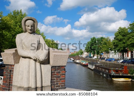 There are the calm street with parking cars, green trees, not high houses and canal with houseboats and boats in the Dutch town. In the foreground the stone sculpture of woman is on the bridge.