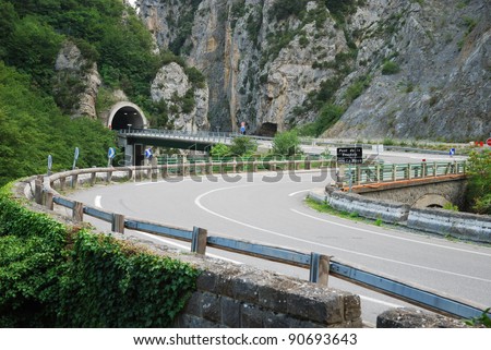 Asphalt road is twisting along sheer cliffs overgrown with lush greenery. Highway is winding through picturesque European Alps in summer.