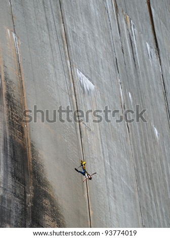 The woman is bungee jumping along the concrete wall. The Contra Dam is a popular bungee jumping venue in the Swiss Alps.
