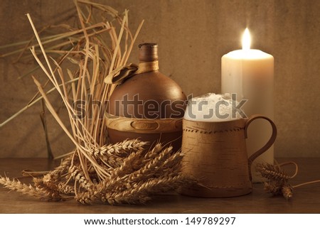 Still life with a clay bottle, wheaten ears a birch bark mug and the foamy drink poured in it and with a candle