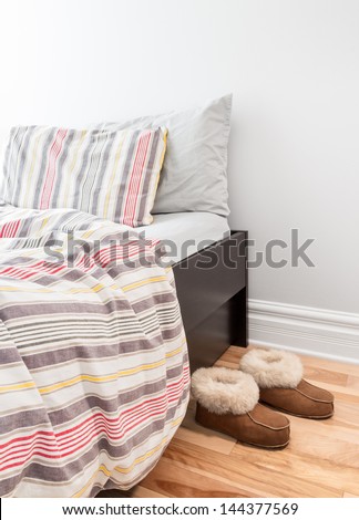 Warm Cozy Slippers Near A Bed With Striped Bed Linen.
