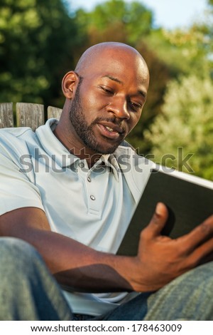 Handsome late 20s black man relaxing reading a book at the park in summer