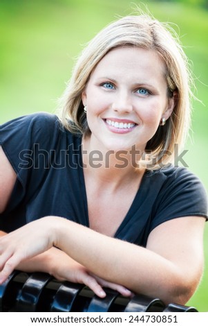 Headshot portrait of an early 30s casual happy beautiful blond woman sitting on a park bench at the park on a sunny summer day