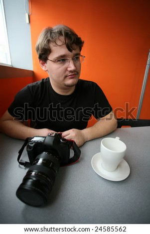 the young photographer sits at the table, on which lies camera and stand coffee cup