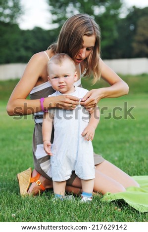 Mom dresses her childboy outdoor on green lawn, he is looking at camera