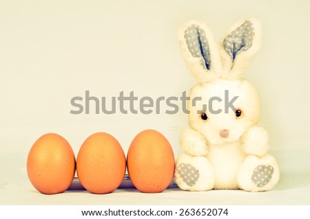 Toy fluffy easter bunny with three orange eggs sits on white background and looking at camera. Eggs are in a row side by side with rabbit.