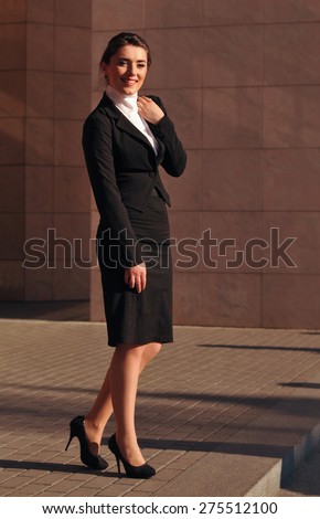 Happy smiling businesswoman full body standing portrait in black suit with skirt and jacket and high-heeled. She smiles and looking at camera.