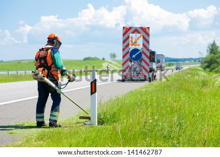 Road landscapers cutting grass around mileposts along the road using string lawn trimmers