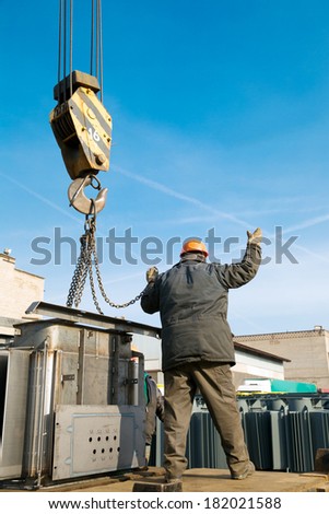 Factory worker gesturing during loading and unloading cargo works with crane at industrial site