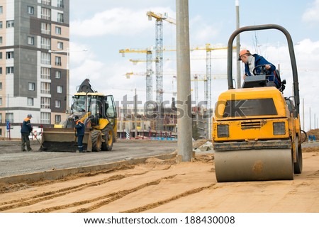 Light tandem vibratory roller compacting sand or soil during construction workers with shovels and tractor vehicle preparation road surface before laying an asphalt driveway