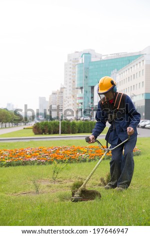 Worker landscaper mowing grass with power tool string lawn trimmer near city building