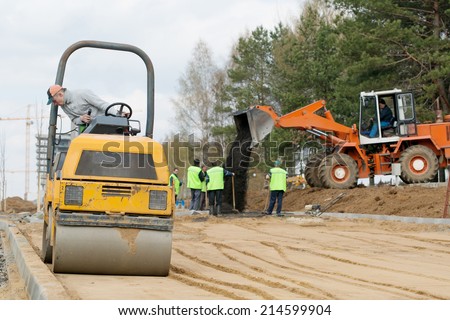 Man worker compacting soil with vibroroller machine along curbstone and wheel loader unloading fresh asphalt during road construction works