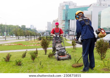 City landscapers gardeners mowing lawn with gas trimmer and lawnmower machine