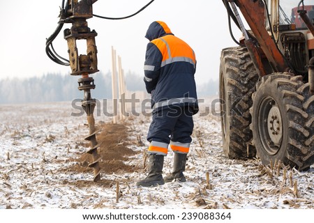 Builder worker monitoring boring holes in ground with drilling rig during fence construction