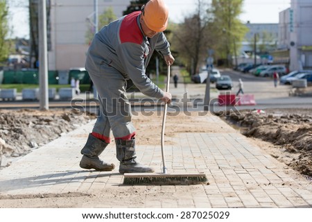 Construction worker grouting dry sand with brush into paver bricks joints during road works