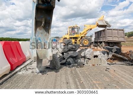 Large jackhammer breaking street asphalt paving during road construction works and wheel loader loading crushing asphalt pieces into dump truck