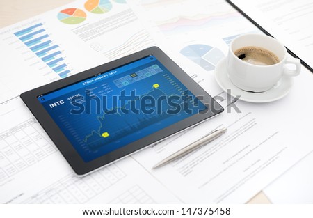 Modern digital tablet with stock market data application on the screen lying on a desk with some papers and documents, pen and a cup of coffee.