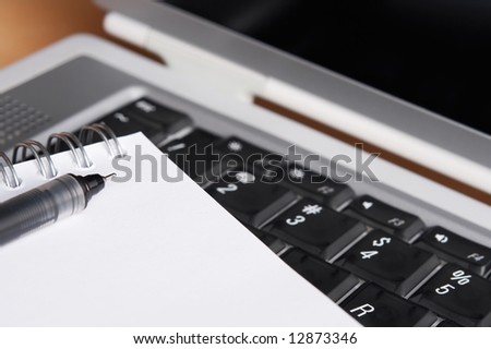 Photo of a laptop, pen and a spiral notebook on a desk. Shallow depth of field. Focus on the pen tip.