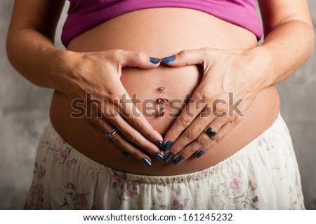 A pregnant woman holding her hands in a heart shape over her baby bump with pierced navel.