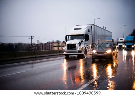 Heavy traffic with big rig semi trucks convoy with semi trailers transporting commercial cargo and another cars on highway in rain evening with headlight reflection on wet surface