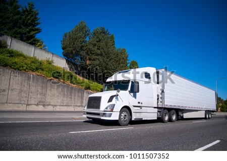 White modern Big rig semi truck with reefer semi trailer equipped with refrigeration unit going on flat wide highway with commercial cargo for delivery to warehouse