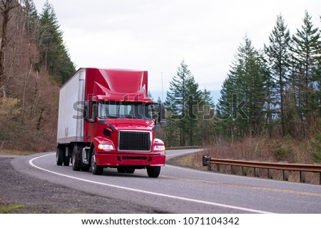 Red big rig day cab semi truck with aerodynamic spoiler on the roof transport dry van semi trailer running on winding road with trees and safety fence on the shoulder
